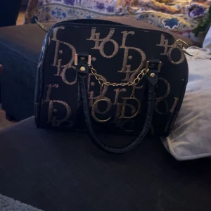 Svart handväska med monogram - Snygg svart handväska med ett iögonfallande monogrammönster i guld. Väskan har två handtag och en kedjedetalj framtill. Perfekt för att ge en lyxig touch till din outfit.