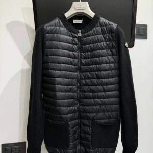 Moncler Cardigan äkta  - Snygg svart cardigan jacka från Moncler med quiltad framsida och stickade ärmar. Jackan har en dragkedja framtill och två praktiska fickor. Perfekt för kyligare dagar med sin stilrena design och Moncler-logga på ärmen. 