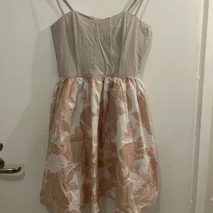 Beige och blommig klänning från H&M - Säljer en söt klänning från H&M med beige korsett överdel och blommigt mönster i rosa och vitt på kjolen. Klänningen har smala axelband och dragkedja bak. 