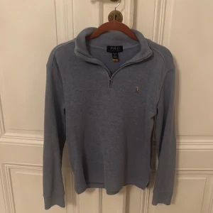 Polo Ralph Lauren half-zip - Säljer en stilren ljusblå tröja från Polo Ralph Lauren med dragkedja och broderad logga på bröstet. Tröjan är i fint skick utan några defekter och är i storlek L/14-16