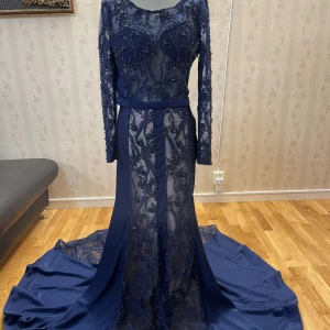 Marinblå långklänning med pärlor - Elegant marinblå långklänning med vackra pärldetaljer och lång ärm. Klänningen har en slits och ett bälte i midjan som ger en smickrande silhuett. Perfekt för formella tillfällen.