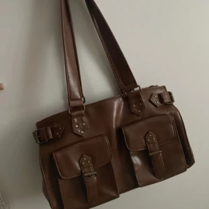 Brun axelväska från Dixie - Snygg brun axelväska från Dixie i skinn med två framfickor och dekorativa spännen. Väskan har dubbla axelremmar och nitar som detaljer.   OBS! Handtagen har lossat lite där de fäster i väskan, jag har limmat de så att de sitter fast men därav reducerade jag priset från 350kr till 300kr! 🫶🏼Kolla gärna in mina andra produkter och testa pruta om du inte är nöjd med priset pga defekterna!