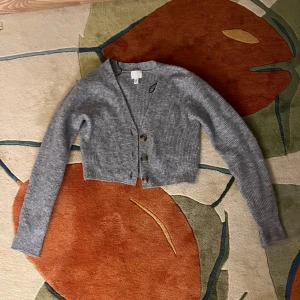 Grå ribbad kofta från H&M - Säljer en  grå ribbad kofta från H&M. Den är croppad med knappar framtill och har långa ärmar. Perfekt för lager-på-lager-stil. Passar till både jeans och klänning!