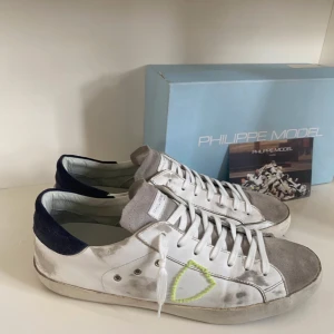 Vita sneakers från Philippe Model - Snygga vita sneakers från Philippe Model med grå detaljer och en unik gul broderad detalj på sidan. Skorna har en klassisk snörning och en mörkblå häl. Perfekta för en stilren look.