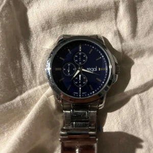 Silverfärgad klocka med blå urtavla från Regal - Snygg klocka från Regal med en stilren blå urtavla och tre mindre urtavlor för extra funktionalitet. Boetten och armbandet är i silverfärgad metall, vilket ger en elegant look. Perfekt för den som vill ha en tidlös accessoar.