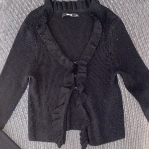 Svart kofta från BikBok - Säljer en svart kofta från BikBok i storlek XS. Koftan har en v-ringad design med volangdetaljer längs kanten och knäppning framtill. Perfekt för att lägga till en touch av elegans till din outfit.