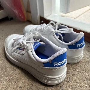 Vita sneakers från Reebok - Snygga vita Reebok sneakers med blå detaljer och klassisk snörning. Perfekta för en stilren look. Skorna har en bekväm passform och är gjorda i syntetmaterial. Dessa skor har använts en gång då dom är för stora för mig, de är i storlek 38,5 och passar perfekt till både tjejer och killar! Nypris är 800kr