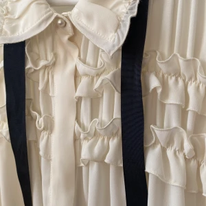 Vit volangblus med svart band från Zara - Benvit volangblus från Zara med ett svart band som kan knytas som man vill. Dold knäppning fram och pärlor vid är slut och närmast kragen. Längd 61. Storlek L. 