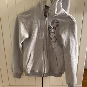 Grå huvtröja med dragkedja från Vonstudio - Säljer en grå huvtröja från Vonstudio i storlek XS. Tröjan har en dragkedja framtill och en stilren design med ett tryck på framsidan. Perfekt för en avslappnad stil.