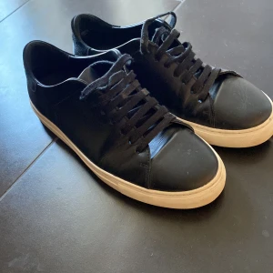 Svarta sneakers från Axel Arigato - Snygga svarta sneakers från Axel Arigato med vit sula och klassisk snörning. Skorna har en stilren design med märkets logga diskret placerad på sidan och sulan. Perfekta för en casual look.