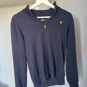 Mörkblå tröja med dragkedja från Lyle & Scott - Snygg mörkblå tröja från Lyle & Scott med dragkedja vid halsen och deras ikoniska gula logga på bröstet. Perfekt för en stilren look. Tröjan har långa ärmar och en ribbad krage.