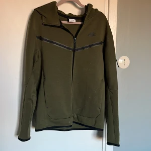 Grön hoodie från Nike - Säljer en grön hoodie från Nike med dragkedja och huva. Den har långa ärmar och en stilren design med svarta detaljer. 