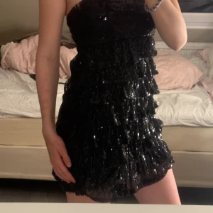 Svart glittrig klänning - Snygg svart klänning med glittriga detaljer och volanger. Perfekt för en festkväll! Klänningen har en kort längd och en tight passform som framhäver figuren.