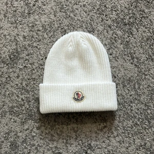Vit ribbad mössa från Moncler - Säljer en stilren vit ribbad mössa från Moncler med deras ikoniska logga framtill. Perfekt för att hålla värmen under kyliga dagar. Passar både till vardags och mer uppklädda tillfällen.