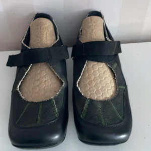 Svarta Boogaloo skor med kardborreband nya - Snygga svarta skor med kardborreband och gröna sömmar. Skorna har en unik design med en fyrkantig tå och kontrasterande ljusa insidor. Perfekta för en stilren look.