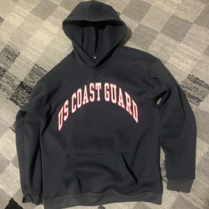 Svart hoodie med US Coast Guard-tryck - Säljer en svart hoodie med texten 'US COAST GUARD' i vitt och rött på bröstet. Tröjan har en klassisk design med huva och långa ärmar. Perfekt för en avslappnad stil.