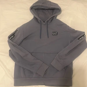 Ljus grå Emporio Armani hoodie - Säljer en stilren grå hoodie från Emporio Armani med EA7-logga på bröstet och textdetaljer på ärmarna. Tröjan har en klassisk huva med dragsko och ribbade muddar vid ärmslut och nederkant. Perfekt för en avslappnad look.