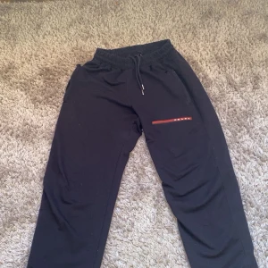 Svarta mjukisbyxor från Prada - Säljer ett par svarta mjukisbyxor från Prada med elastisk midja och snörning. Byxorna har en diskret röd detalj vid fickan som ger en snygg kontrast. Perfekta för en avslappnad stil.