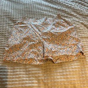 Blommiga shorts från Name It - Säljer ett par söta blommiga shorts från Name It. De har ett färgglatt mönster med lila och orange blommor på en vit bakgrund. Perfekta för varma sommardagar med en bekväm passform och elastisk midja, använda en gång 