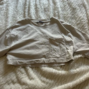 Beige croppad topp från Zara - Säljer en beige croppad topp från Zara med en bröstficka. Toppen har korta ärmar och en rund halsringning. Perfekt för en avslappnad stil.