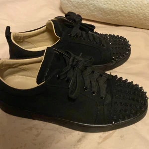 Christian Louboutin  -  TAR HELST BYTEN Snygga svarta sneakers med nitar på tån och svarta snören. Skorna har en röd sula som ger en cool kontrast. Perfekta för att ge din outfit en edgy touch.
