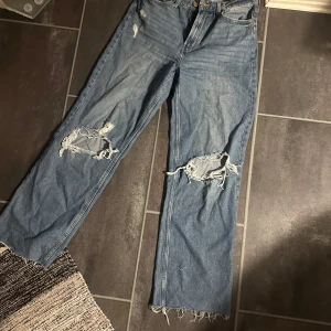 Blå jeans med slitningar - Säljer ett par blå jeans från &Denim med coola slitningar på knäna och fransiga kanter. De har en ultra hög midja och en bred passform som ger en avslappnad look. Perfekta för en trendig stil!