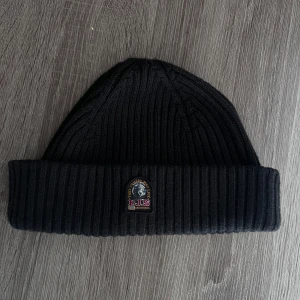 Svart ribbad mössa från Parajumpers - Snygg svart ribbad mössa från Parajumpers med ett broderat märke framtill. Perfekt för att hålla värmen under kyliga dagar. Stilren och enkel design som passar till det mesta.