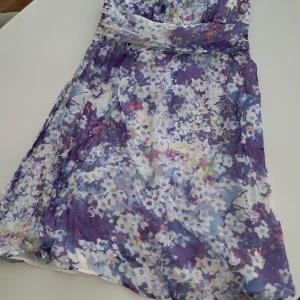 Blommig sidenklänning från Une Deux Trois - Säljer en vacker blommig klänning i SIDEN från Une Deux Trois. Klänningen har en härlig mix av lila, blå och vita toner med ett blommigt mönster. Den är axelbandslös och har en dragkedja i ryggen. Perfekt för sommarens festligheter!