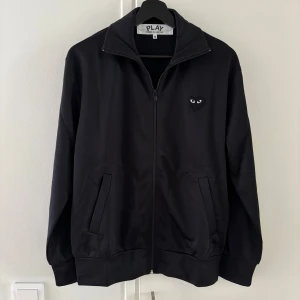 Svart Comme des Garçons zip up - Säljer en stilren svart zip up tröja från Comme des Garçons med det ikoniska hjärtmotivet med ögon på bröstet och ryggen. Den har en dragkedja och två fickor framtill. Är i storlek M men passar även S.