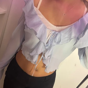 Ljusblå blus med volangdetaljer - Säljer en ljusblå blus med volangdetaljer och knytning framtill. Blusen har långa ärmar och en luftig passform, perfekt för vår och sommar. Den är tillverkad i ett lätt och skönt material.