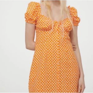 Orange blommig klänning med puffärmar - Säljer en söt orange klänning med vita blommor och puffiga kortärmar. Klänningen har en smickrande passform och är perfekt för sommarens varma dagar. Den har en v-ringad design som ger en feminin touch.
