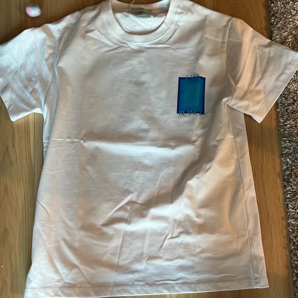 Säljer en stilren vit t-shirt från Casablanca med ett blått grafiskt tryck på både fram- och baksidan. T-shirten har en klassisk rund hals och korta ärmar. Perfekt för en avslappnad look!. T-paidat.