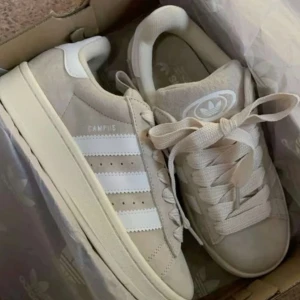 Beige Adidas Campus sneakers mocka - Snygga Adidas Campus sneakers i beige mocka med klassiska vita ränder på sidorna och breda snören. Skorna har rund tå, platt sula och Adidas logga på plösen. Perfekta för dig som gillar stilrena och trendiga sneakers med retrovibbar.