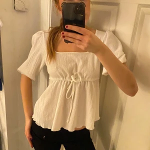 Vit blus med knytband - Säljer en söt vit blus med korta ärmar och knytband i midjan. Blusen har en lätt ribbad struktur och en luftig passform, perfekt för varmare dagar.