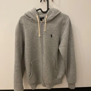 Grå hoodie från Ralph Lauren - Säljer en stilren grå hoodie från Ralph Lauren med dragkedja och klassisk logga på bröstet. Den är använd men i fint skick, hör av dig vid fler frågor!