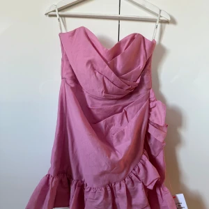 Rosa klänning från ASOS DESIGN - Säljer en söt rosa klänning från ASOS DESIGN. Klänningen har en volangdetalj och är axelbandslös, perfekt för sommarens fester. Tillverkad i ett lätt och bekvämt material. Helt oanvänd med lappen kvar. 
