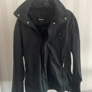 Svart vindjacka från Bench - Snygg svart vindjacka från Bench med dragkedja och hög krage. Jackan har långa ärmar och Bench-logga på kragen. Perfekt för blåsiga dagar.
