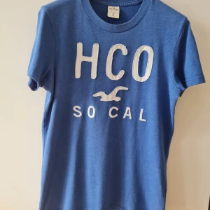 Blå t-shirt från Hollister - Säljer en blå t-shirt från Hollister med vit text HCO SO CAL och hollister fågeln på framsidan. Den är kortärmad och gjord i mjukt bomullsmaterial. Storlek S. Kan hämtas i Vallentuna.