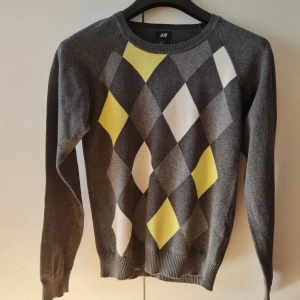Grå tröja med diamantmönster från H&M - Säljer en stilren grå tröja från H&M med ett coolt diamantmönster i gult, vitt och olika grå nyanser på framsidan. Tröjan har långa ärmar och ribbade kanter vid ärmslut och nederkant. 100% bomull. Storlek 170. Kan hämtas i Vallentuna.
