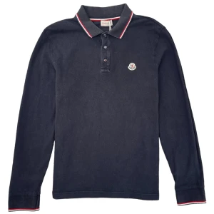 Svart långärmad pikétröja från Moncler - Snygg svart långärmad pikétröja från Moncler med klassisk krage och knappar framtill. Tröjan har diskreta röda och vita detaljer på kragen och ett broderat Moncler-emblem på bröstet. Perfekt för en stilren look. Endast Intressekoll! Storlek L men sitter som m!👍🏼