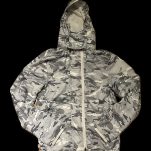 Windbreaker Superdry - Säljer en grå camouflagemönstrad vindjacka från Superdry. Jackan har en huva och dragkedja framtill. Perfekt för blåsiga dagar med sin stilrena design och praktiska fickor. Superdry-logga på ryggen och armen ger en extra touch. Storleken är S men passar mig på 185cm som brukar ha M-L. Den har 3 lager med dragkedjor så man kan välja själv hur tajt den ska sitta. Har även spännen längst ner man kan dra åt.