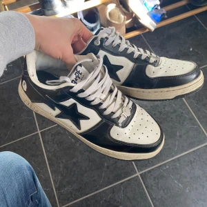 Svartvita sneakers från Bape - Säljer ett par stilrena svartvita sneakers från Bape. Skorna har en klassisk design med en svart stjärna på sidan och vita snören. Perfekta för en casual look.