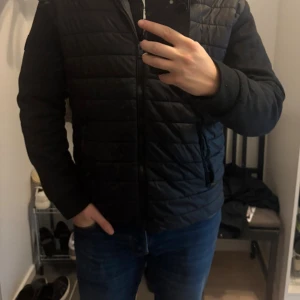 Svart quiltad pufferjacka med huva Lindbergh - Svart pufferjacka lindbergh med quiltad framsida och huva. Jackan har dragkedja framtill, två fickor med dragkedja och släta ärmar. Perfekt för kyliga dagar och har en clean, stilren look. Storlek XL