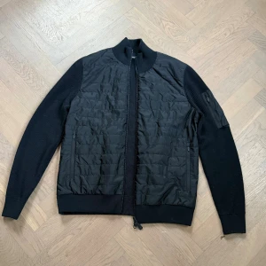 Svart cardigan jacka från J.Lindeberg - Snygg svart cardigan jacka från J.Lindeberg med quiltad framsida och stickade ärmar och rygg. Knappast använd