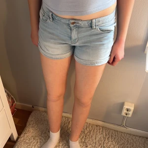Ljusblå jeansshorts från Lindex - Säljer ett par ljusblå jeansshorts från Lindex med klassisk femficksdesign och uppvikta benslut. Shortsen har normal passform, bälteshällor och stängs med knapp och dragkedja. 