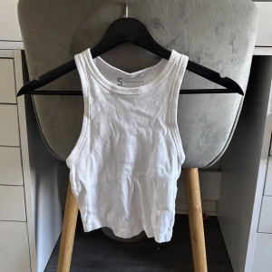 Vit ribbad linnetopp i bomull - Säljer en vit ribbad linnetopp i bomull med rund halsringning och ärmlös design. Perfekt för varma dagar och enkel att matcha med jeans eller kjol. Snygg croppad passform som ger en trendig look.