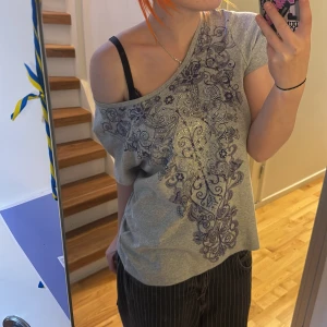 Grå topp med lila mönster från Zenergy - Säljer en grå topp från Zenergy by Chico's med snyggt lila, blommigt och paisleyinspirerat tryck framtill. T-shirten har korta ärmar och är i mjuk bomullsblandning. 