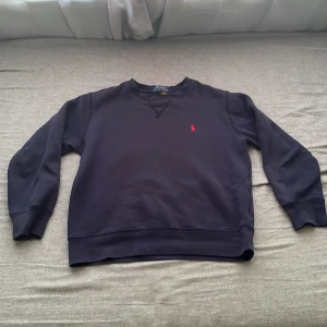 Mörkblå sweatshirt från Polo Ralph Lauren - Mörkblå sweatshirt från Polo Ralph Lauren med klassisk rund hals och röd broderad logga på bröstet. Tröjan har långa ärmar och ribbade muddar vid ärmslut och nederkant. Perfekt för en chill och stilren look. Funkar utmärkt till hösten då den håller värmen mycket bra, hör av dig vid frågor mvh Alex 