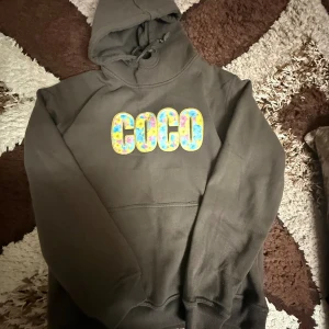 Svart hoodie med färgglad COCO-text - Cool svart hoodie med stor färgglad COCO-text framtill. Tröjan har huva med snörning och en klassisk känguruficka. Perfekt för dig som gillar streetstyle och vill sticka ut lite extra.