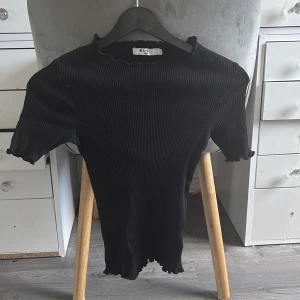 Svart ribbad topp från NA-KD - Svart ribbad topp från NA-KD med korta ärmar och volangkant vid hals, ärmslut och nederkant. Toppen har en tight passform och är tillverkad i stretchigt material som sitter snyggt på kroppen. Perfekt att styla med jeans eller kjol.